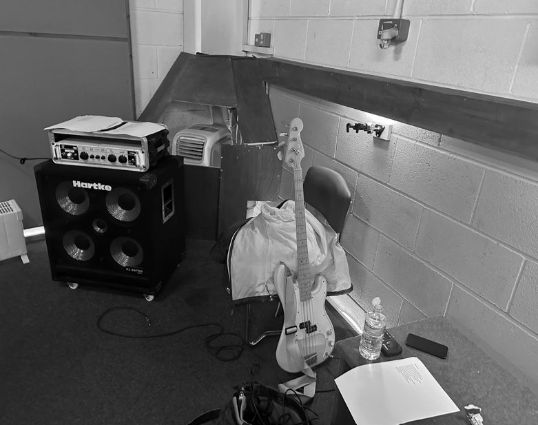 Black and white photo of a Hartke bass amp and cabinet with a bass guitar resting on a chair in a rehearsal room